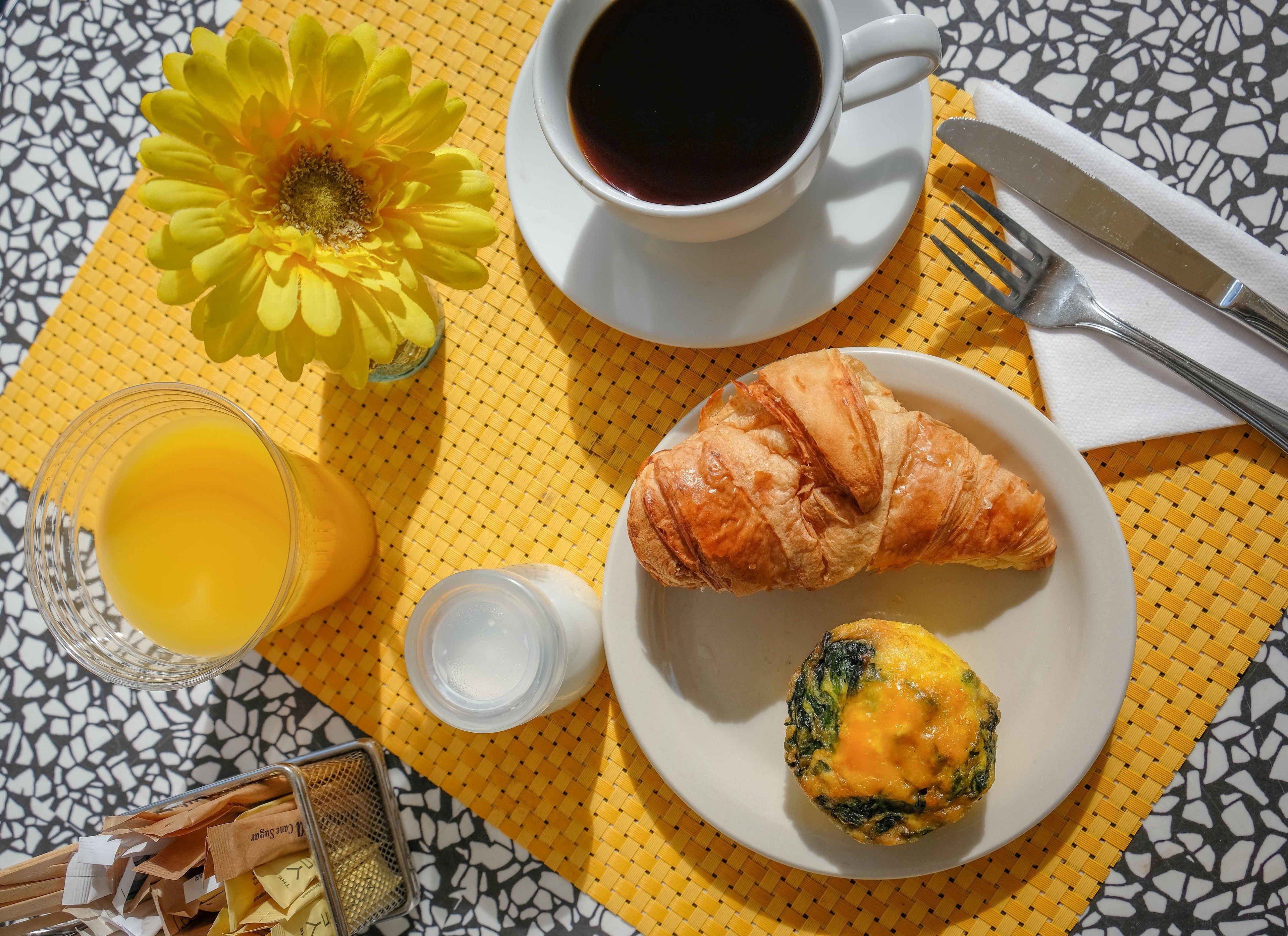 A breakfast setting with a croissant, muffin, coffee, and orange juice on a table with a yellow placemat.