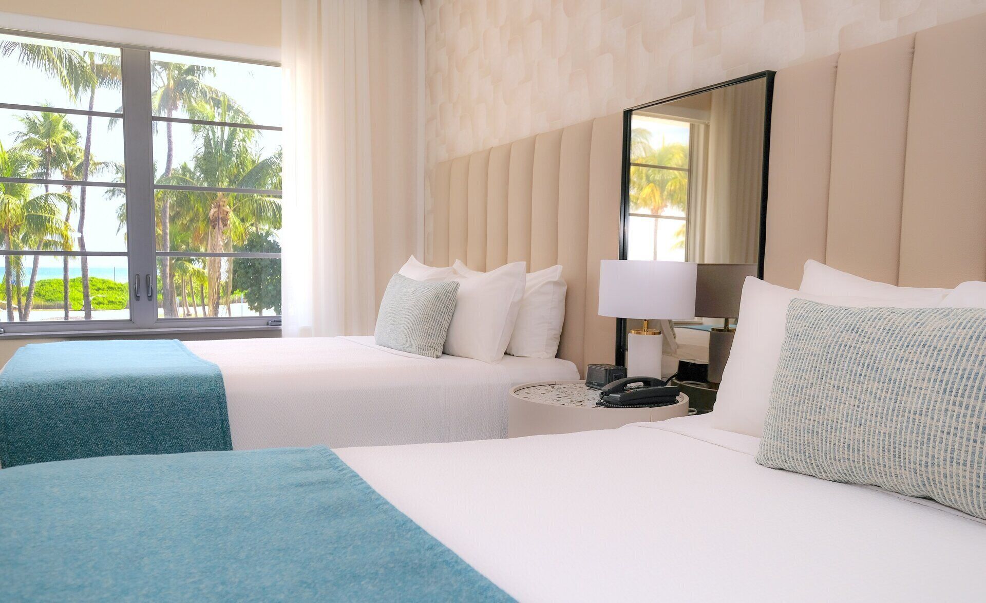 A hotel room with two beds, a large window with a view of palm trees, a bedside table with a lamp, and a mirror on the wall.