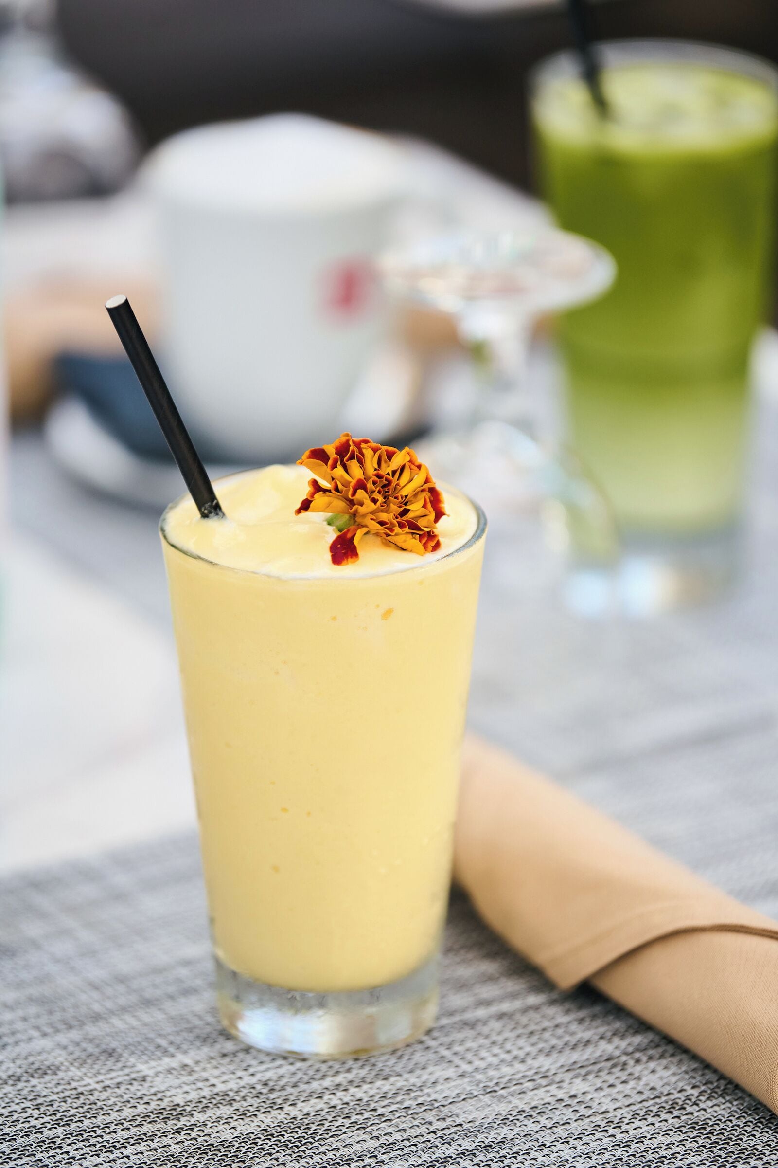 A glass of yellow beverage with a flower garnish and a black straw, placed on a textured surface with a napkin beside it.