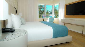Quarto de hotel com uma cama, uma mesa de trabalho com telefone, uma cadeira, televisão e uma janela com vista para palmeiras.