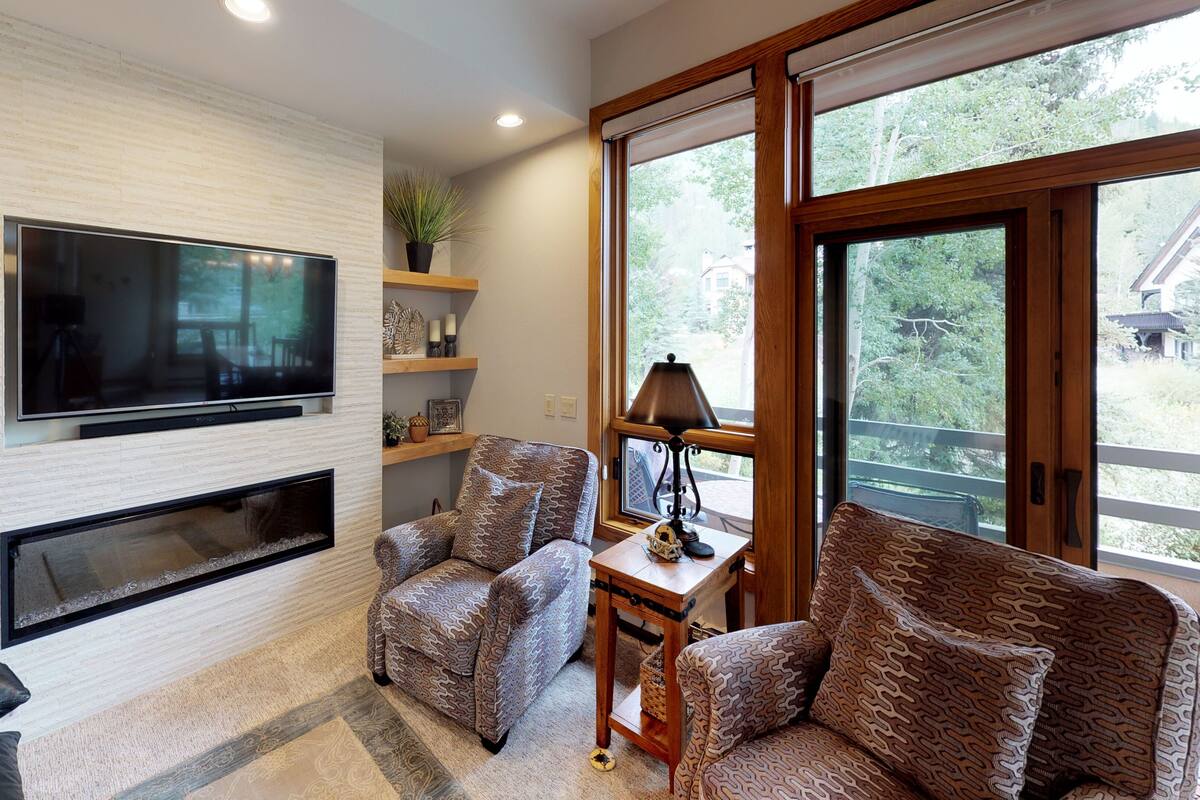 A modern living room with a flat-screen TV mounted on a brick wall, a fireplace, patterned furniture, and a large window offering a view of the outdoors.