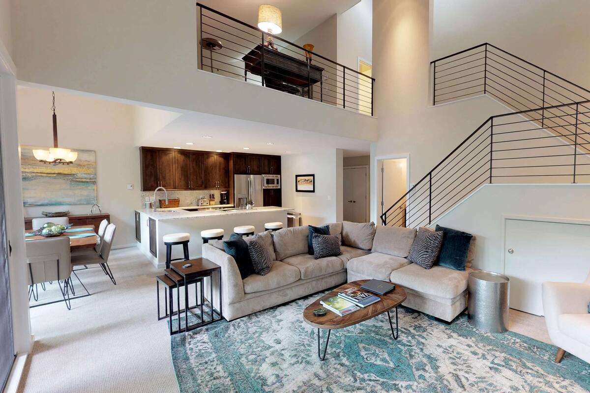 A spacious living area with a large sofa, a coffee table, and a dining table with chairs. There is a kitchen in the background and a staircase leading to an upper level.