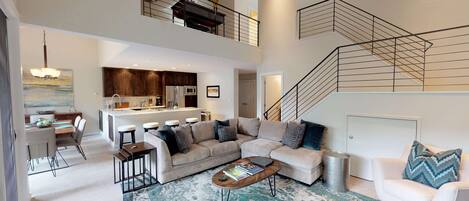 A spacious living area with a large sofa, a coffee table, and a dining table with chairs. There is a kitchen in the background and a staircase leading to an upper level.