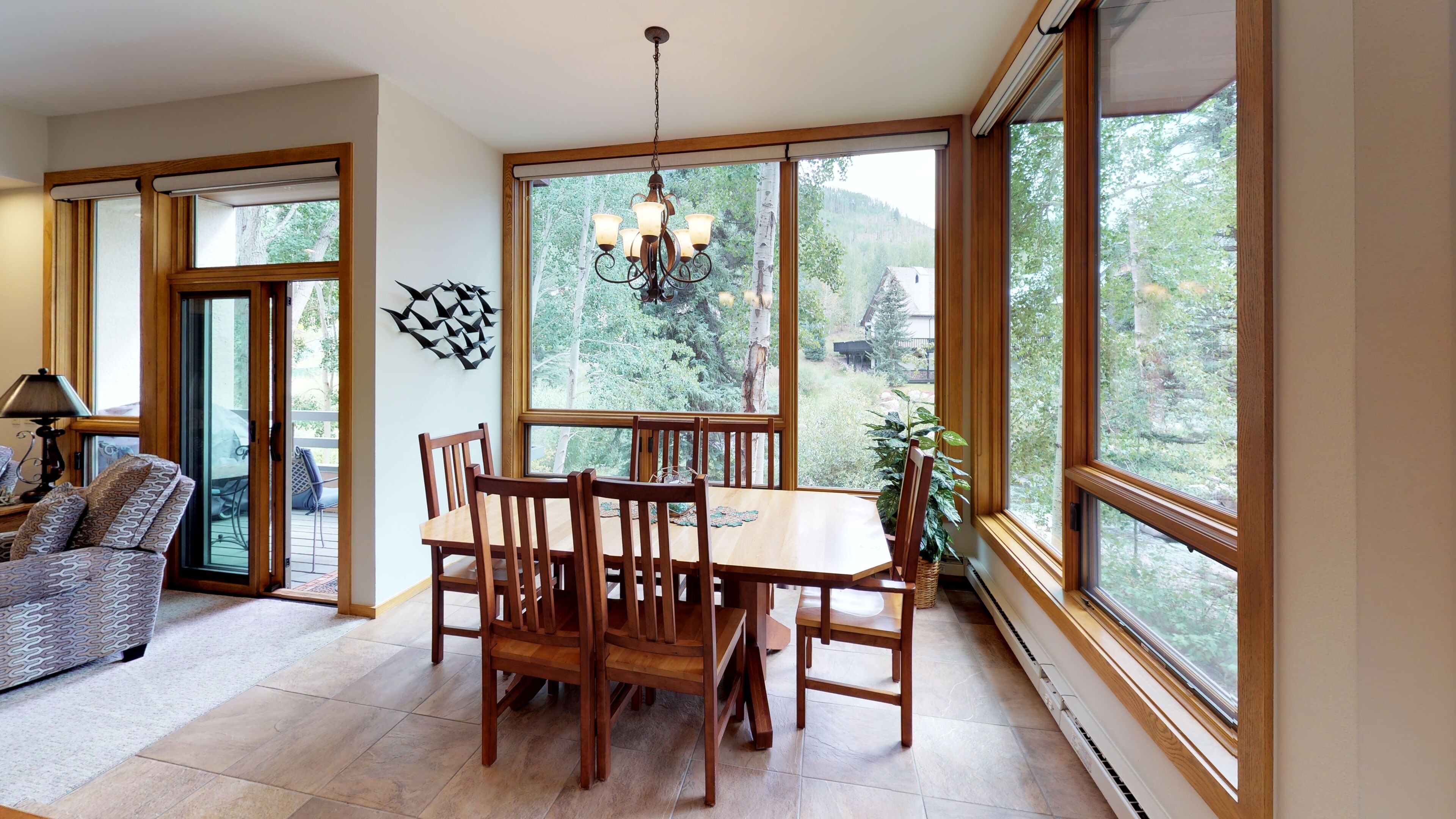 Un comedor con mesa y sillas de madera, ventanas amplias con vista a árboles y una araña.