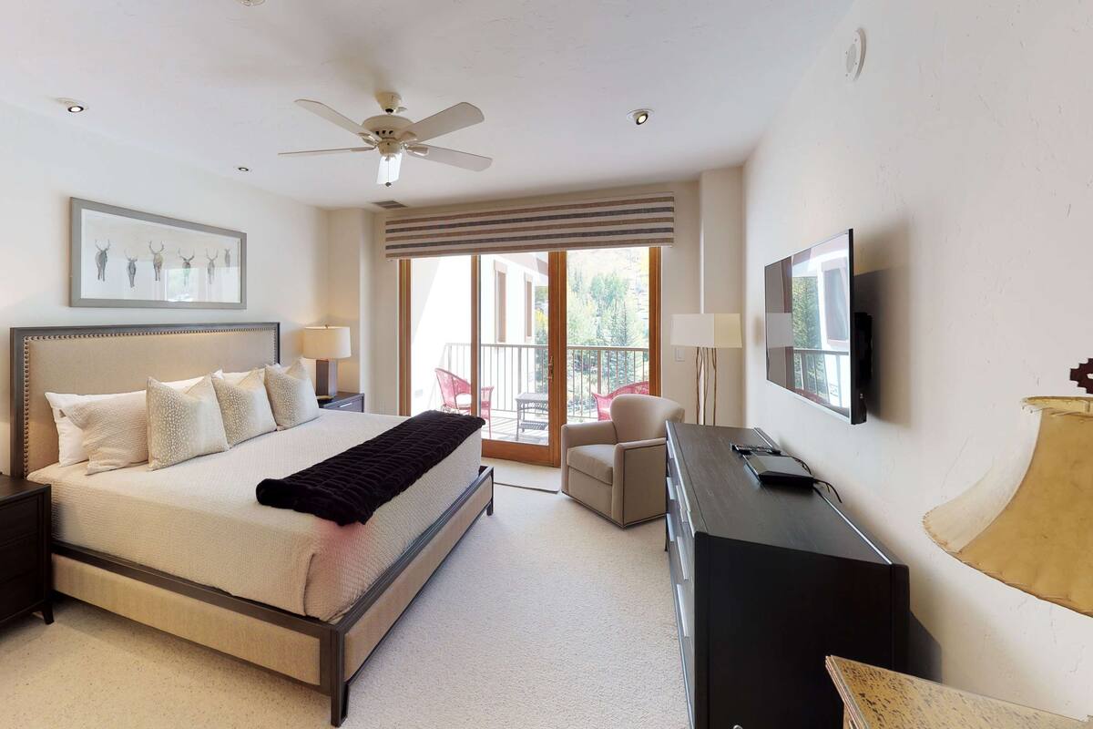 A bedroom with a bed, bedside tables, a desk, a chair, a ceiling fan, a TV, and a balcony with a view.