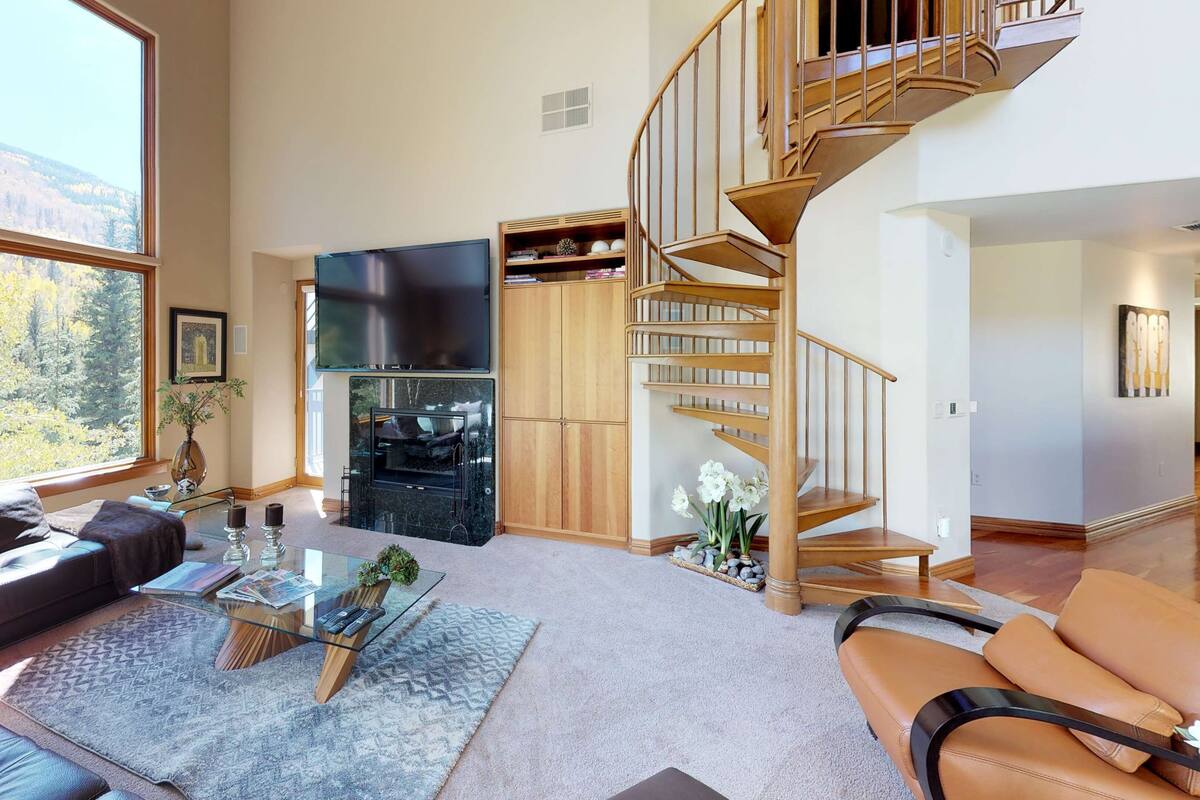 A spacious living room with a fireplace, a large window with a mountain view, a wooden staircase, and a cozy seating area.