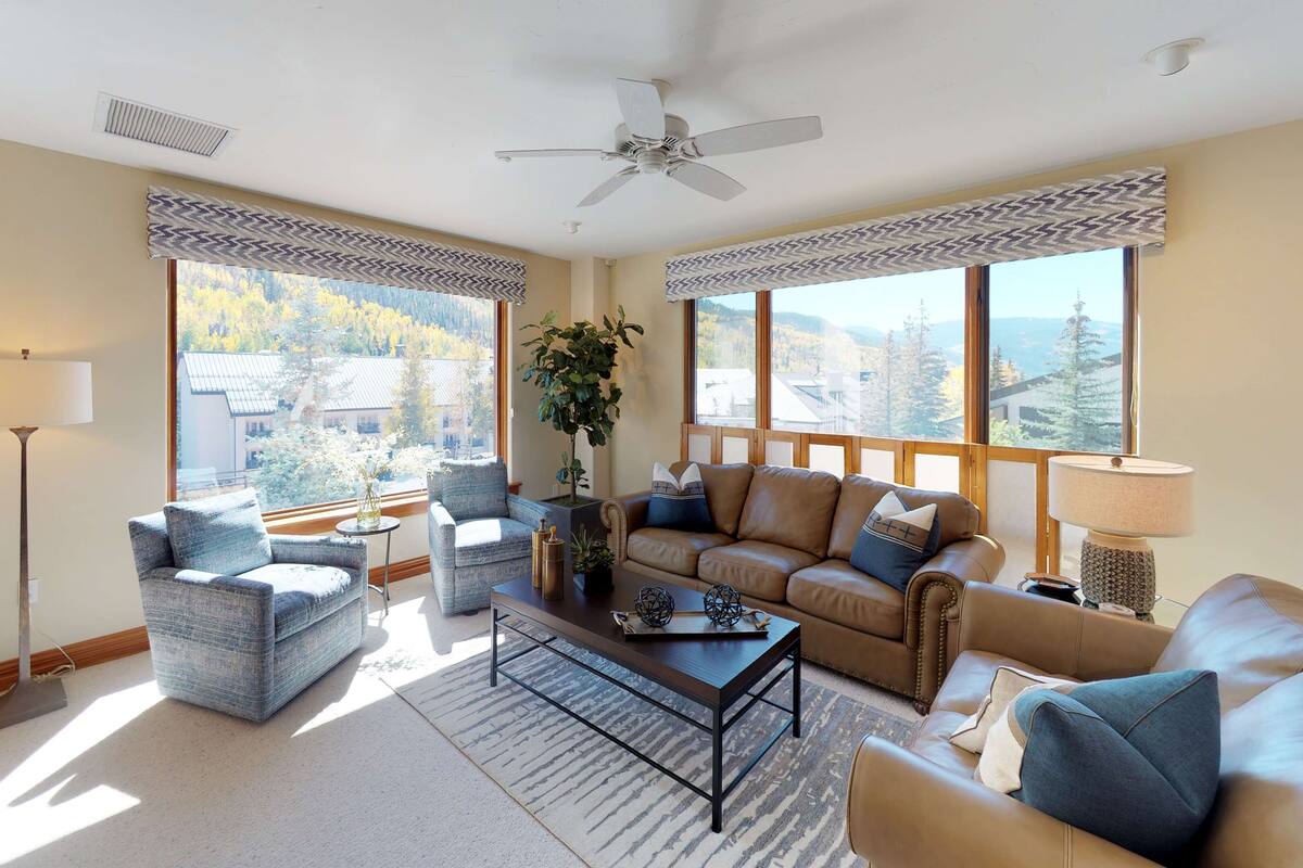 A spacious living room with a high ceiling, large windows offering a view of a mountainous landscape, a ceiling fan, and a mix of seating including a sofa and armchairs.