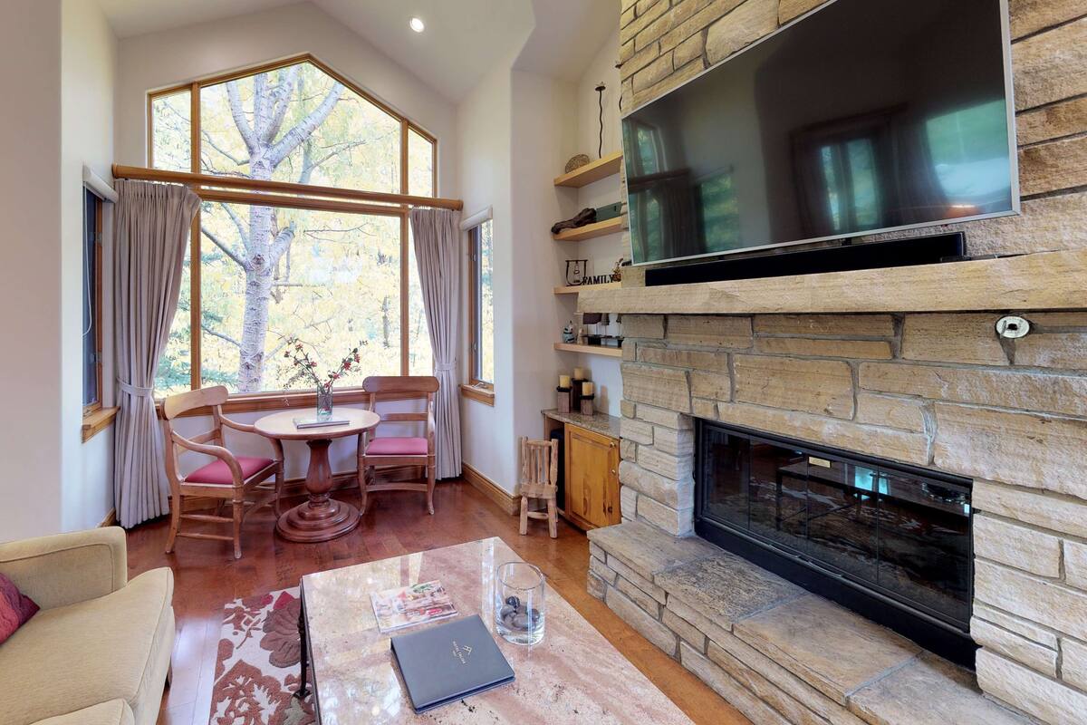 A cozy living room with a fireplace, a sofa, a dining table with chairs, and large windows with curtains.