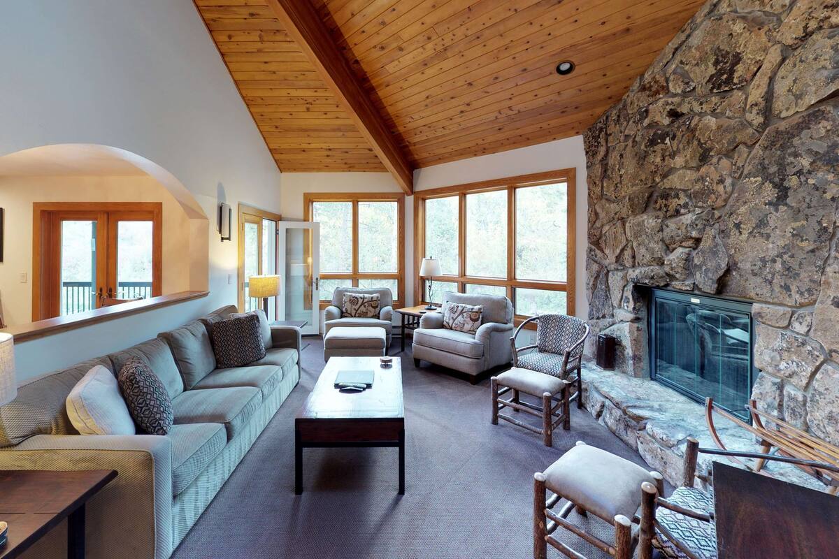A spacious living room with a stone fireplace, wooden ceiling beams, and large windows.
