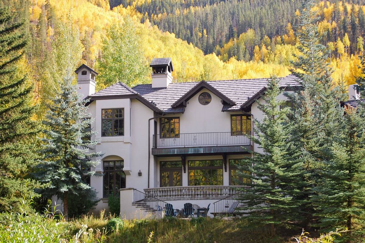 A two-story house with a balcony, surrounded by trees and a mountainous backdrop.