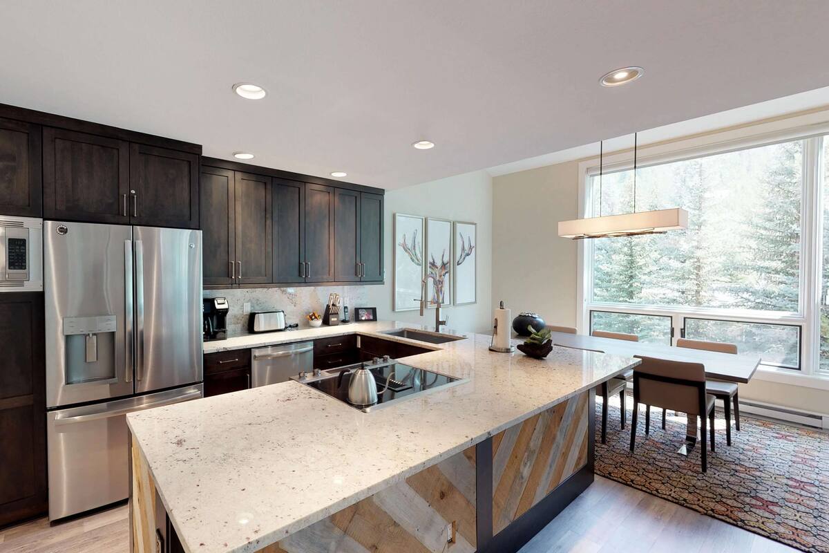 A modern kitchen with dark wood cabinets, stainless steel appliances, a large island with a built-in stove, and a large window providing a view of trees.