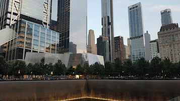 A modern cityscape with high-rise buildings, a large reflective water feature, and a plaza with trees.