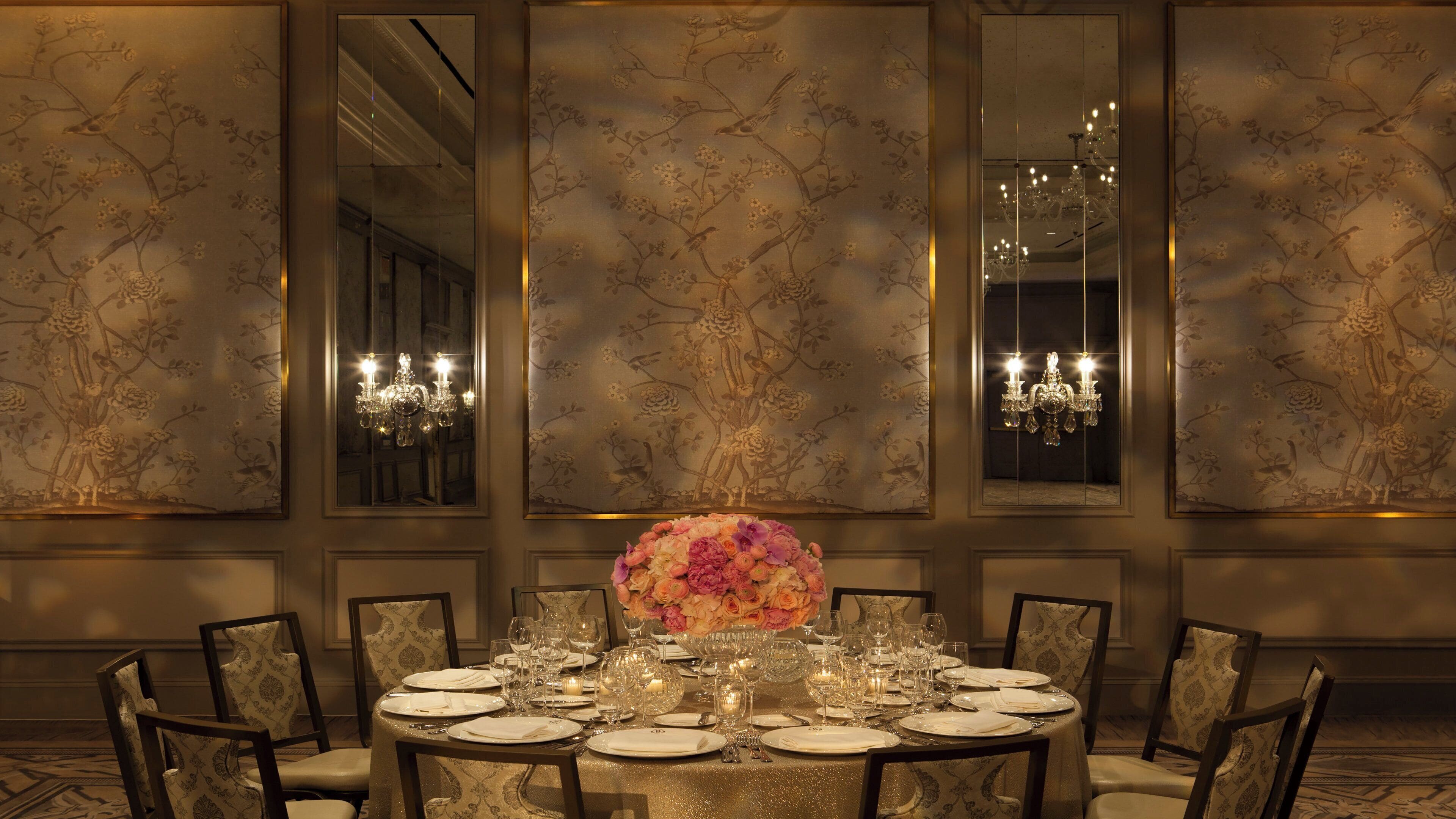 A formal dining room with a round table set for a meal, flanked by chairs, and a large mirror reflecting the room's decor.