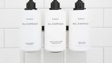 Three white bottles of BYREDO BAL D'AFRIQUE hair products on a white tiled wall.