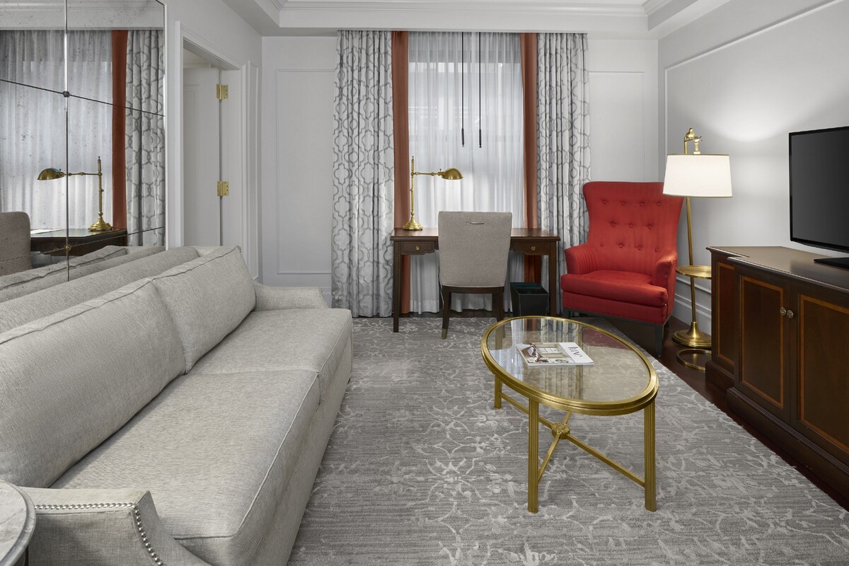A modern hotel room with a sofa, a glass coffee table, a red armchair, a television, and a desk with a lamp.