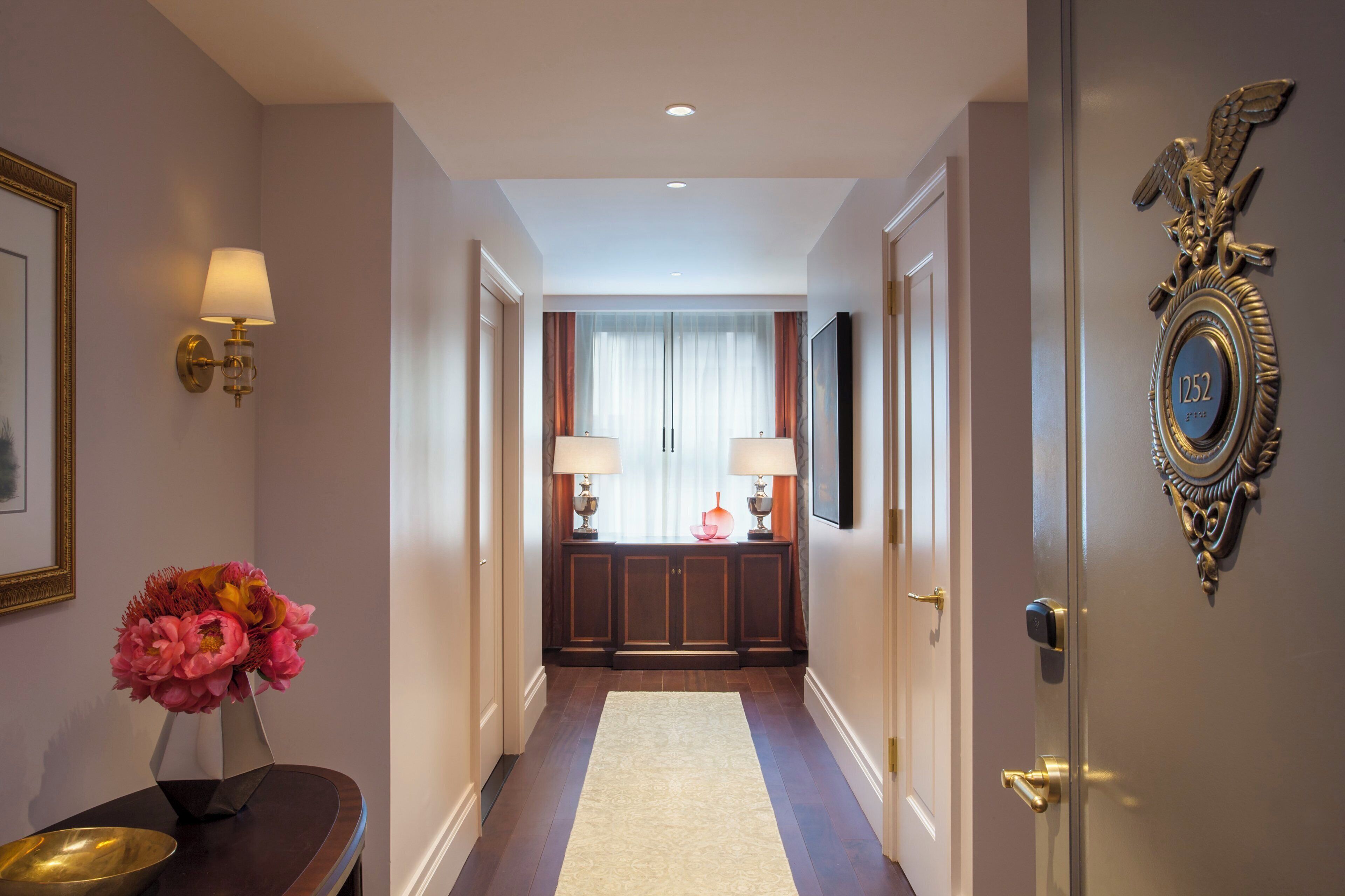 A hotel hallway with a wooden bench, a vase of flowers, wall-mounted lamps, a mirror, and a door with a number plaque.