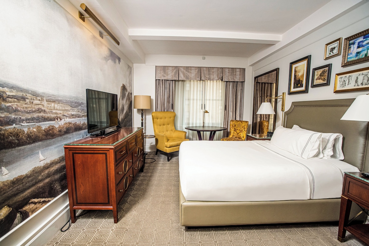 A hotel room with a large bed, a television on a wooden dresser, a yellow armchair, a small round table, and a painting on the wall.