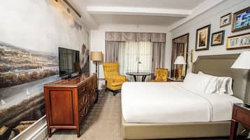A hotel room with a large bed, a television on a wooden dresser, a yellow armchair, a small round table, and a painting on the wall.