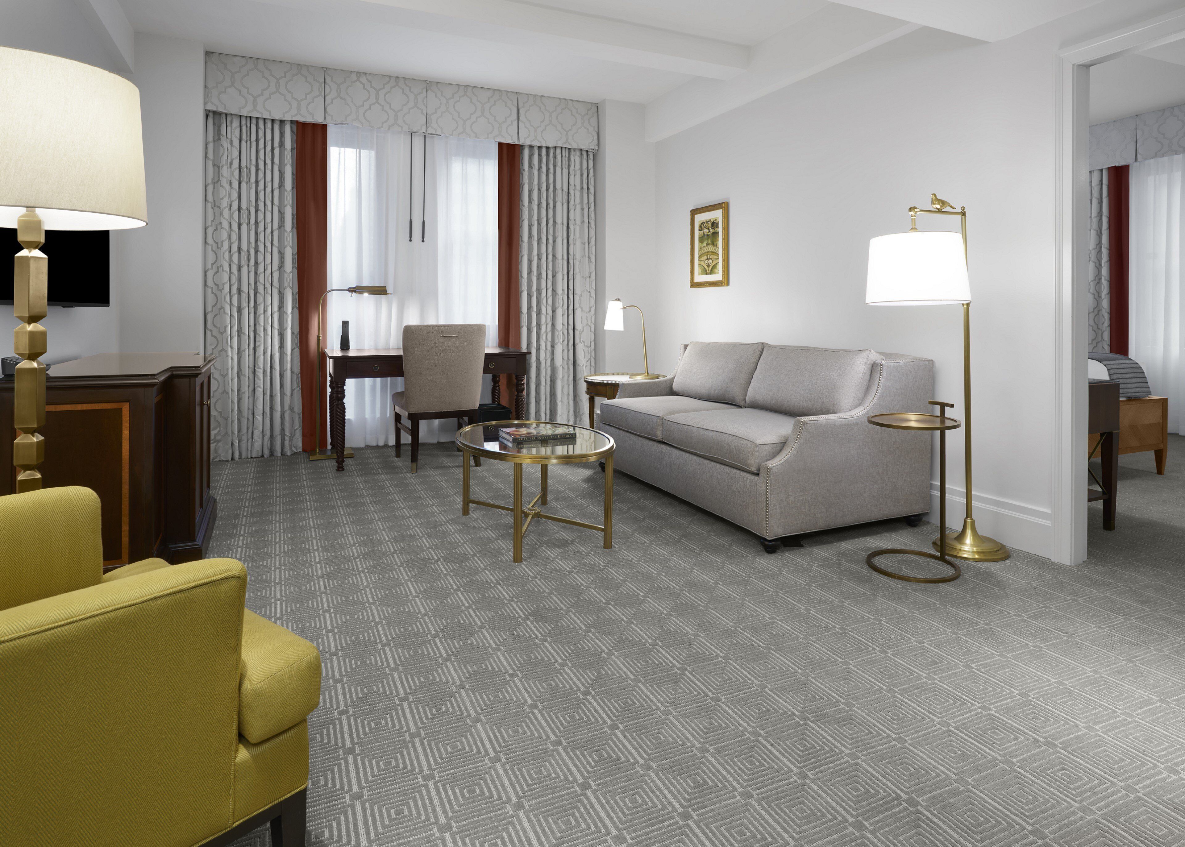 A hotel room with a sofa, armchair, and a small table. There is a desk with a chair and a lamp. The room has patterned carpet and curtains.