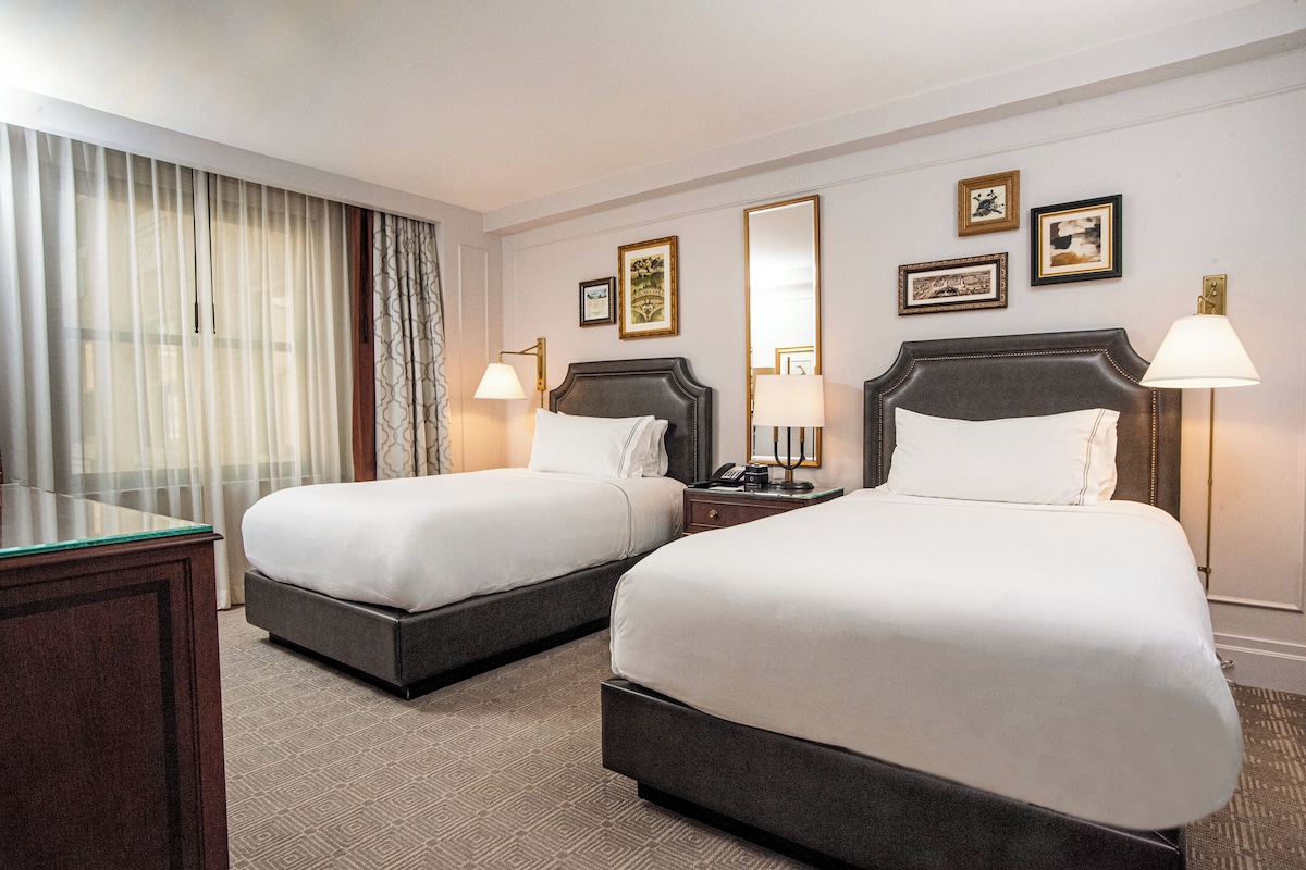A hotel room with two beds, a desk, and a large window with curtains.