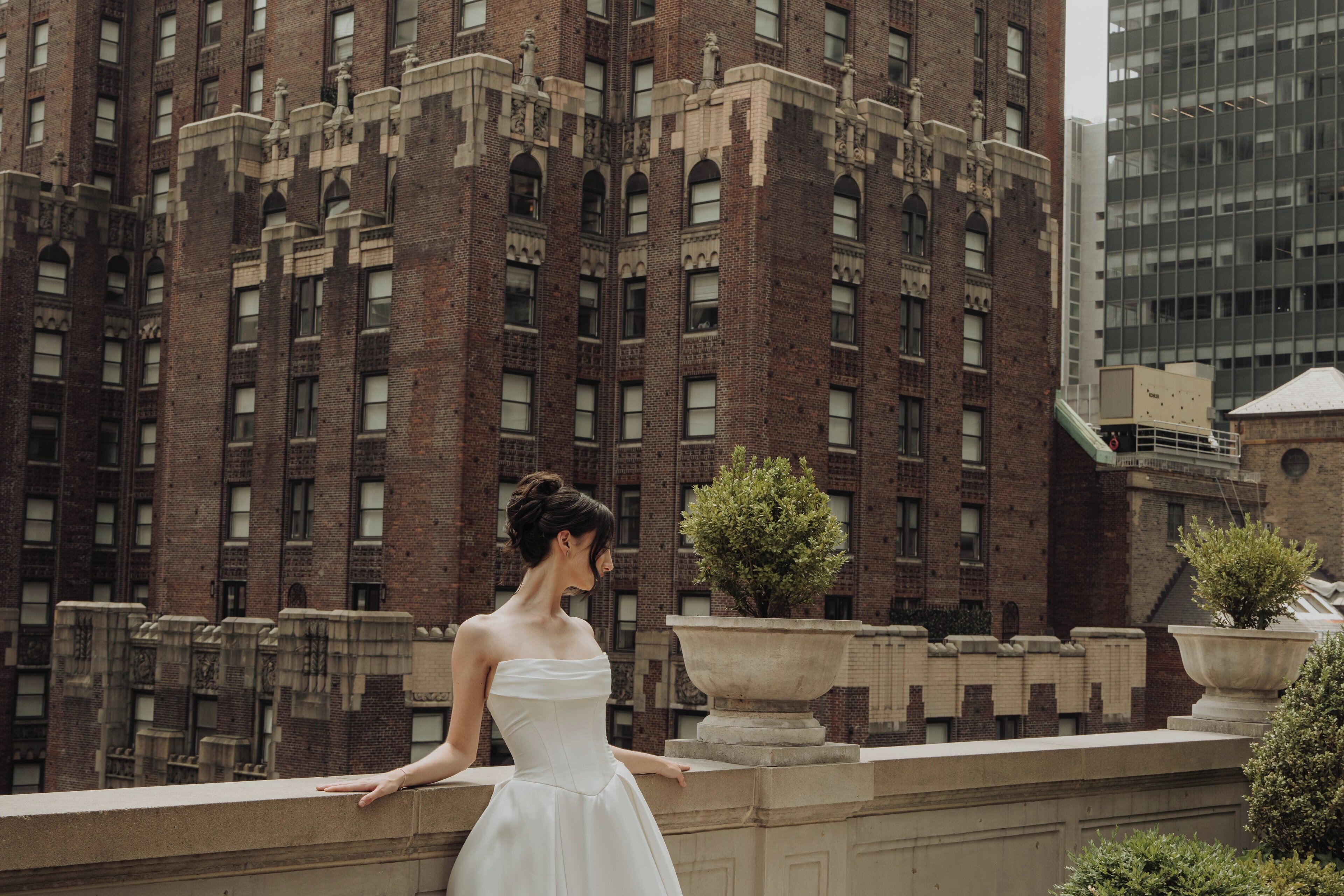 A woman in a white dress standing on a balcony with a view of a large brick building and a modern glass structure in the background.