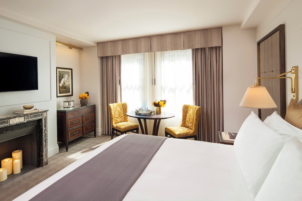 A hotel room with a bed, a TV, a fireplace, a desk, two chairs, a table with flowers, and curtains.