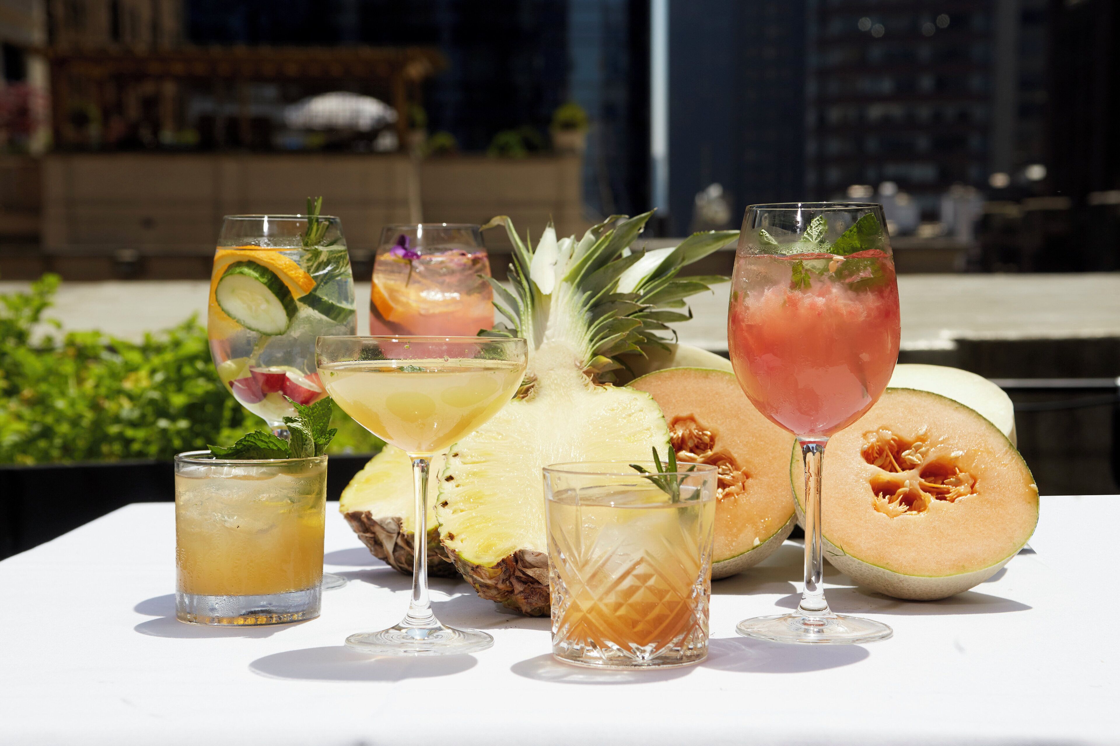 Une table dressée à l’extérieur, sur laquelle on trouve diverses boissons et des fruits, notamment un ananas et un melon.