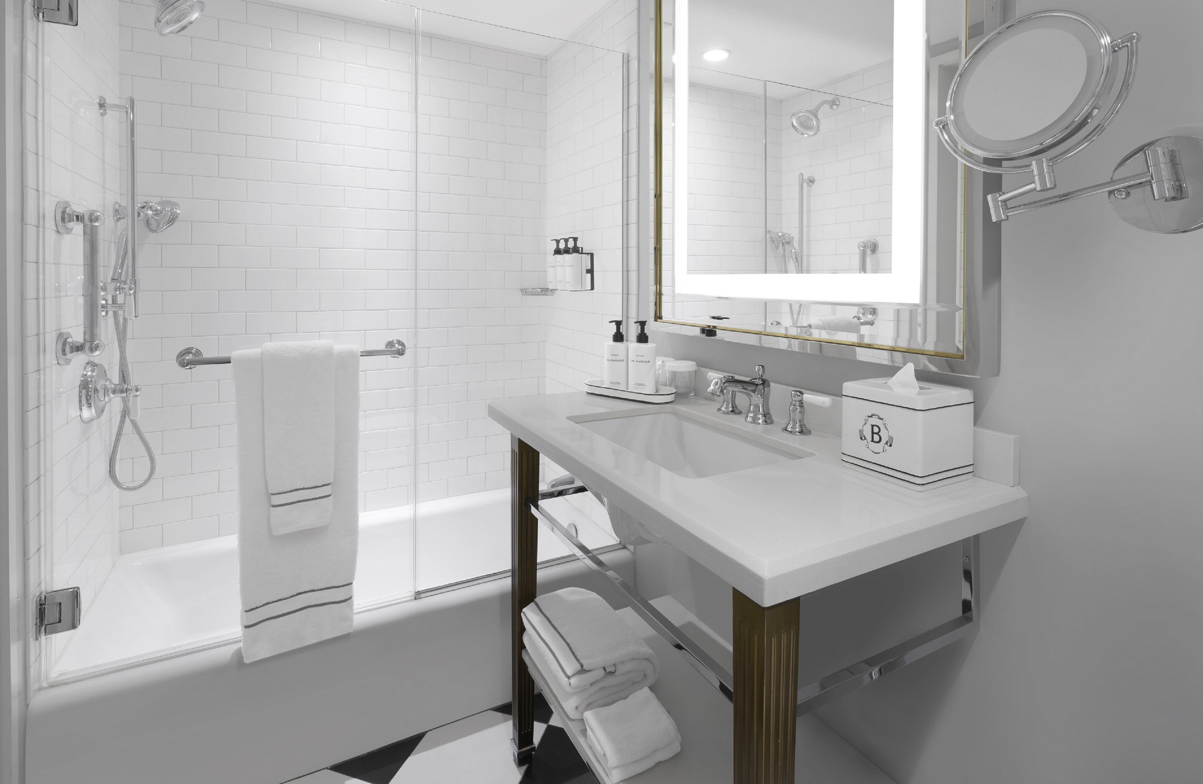 A modern bathroom with a white tiled shower, a large mirror, and a white sink with a gold faucet.