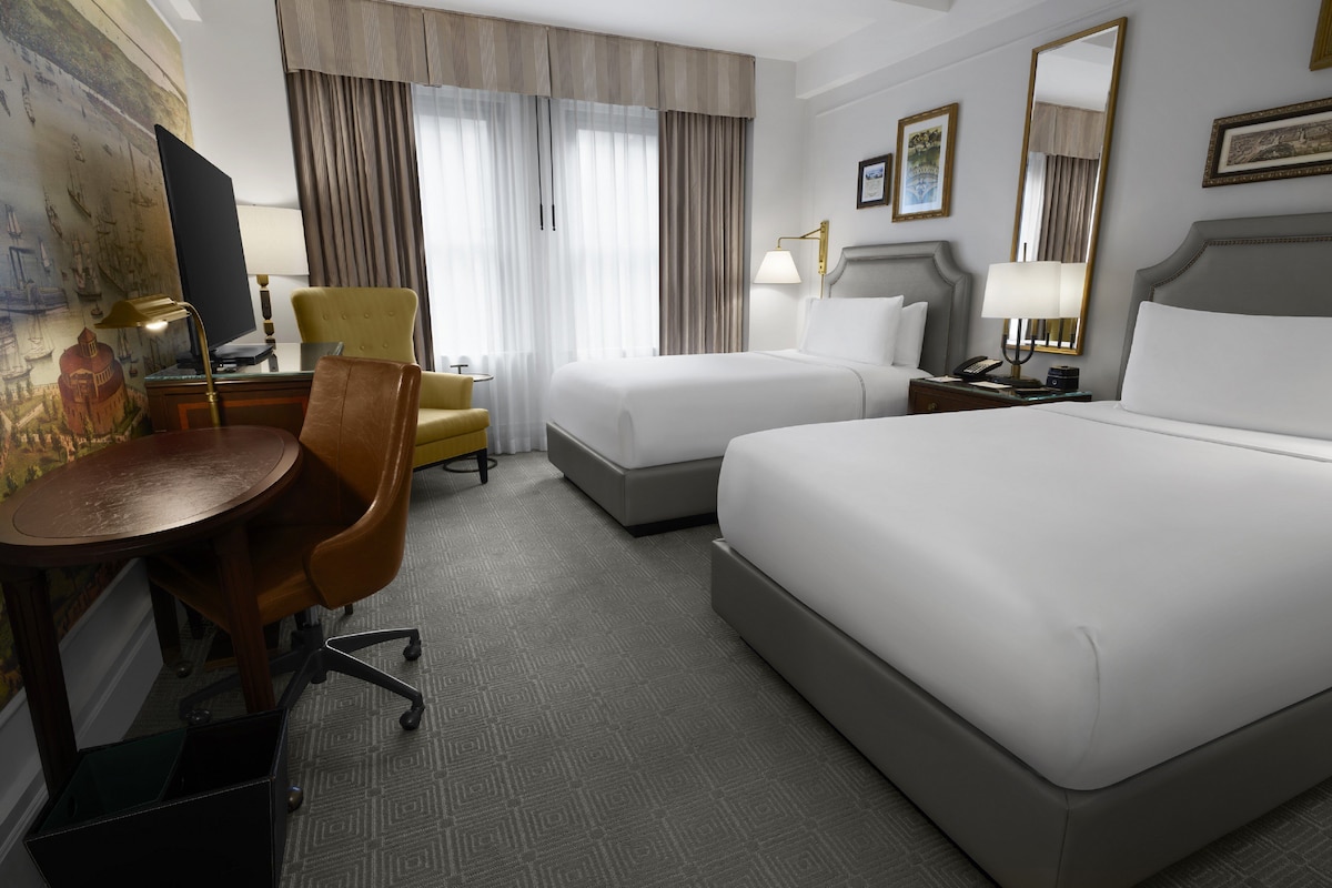 A hotel room with two beds, a desk, a chair, a lamp, and a large mirror.