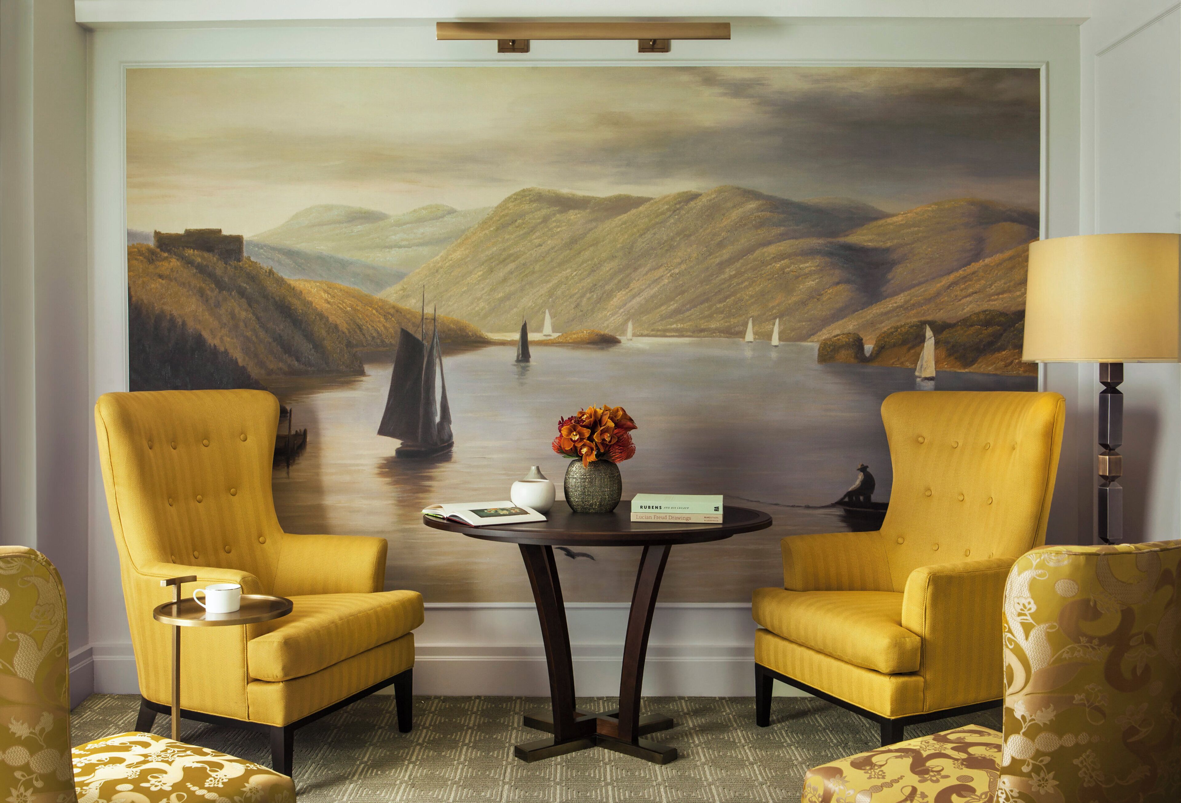 Una habitación con dos sillones amarillos, una mesita con un jarrón de flores y un cuadro grande de paisaje en la pared.