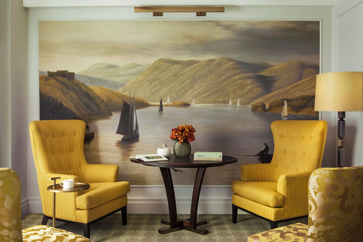 A room with two yellow armchairs, a small round table with a vase of flowers, and a large landscape painting on the wall.