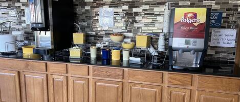 A coffee station with a Folgers coffee maker, a coffee dispenser, and various cups and containers.