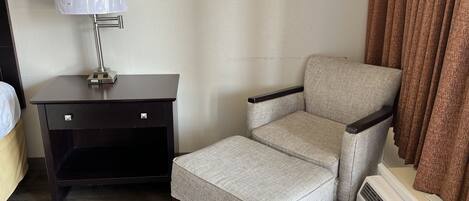 Una habitación de hotel con una mesita de noche de madera oscura, una lámpara, un sillón gris, una pequeña mesa y un equipo de aire acondicionado blanco.
