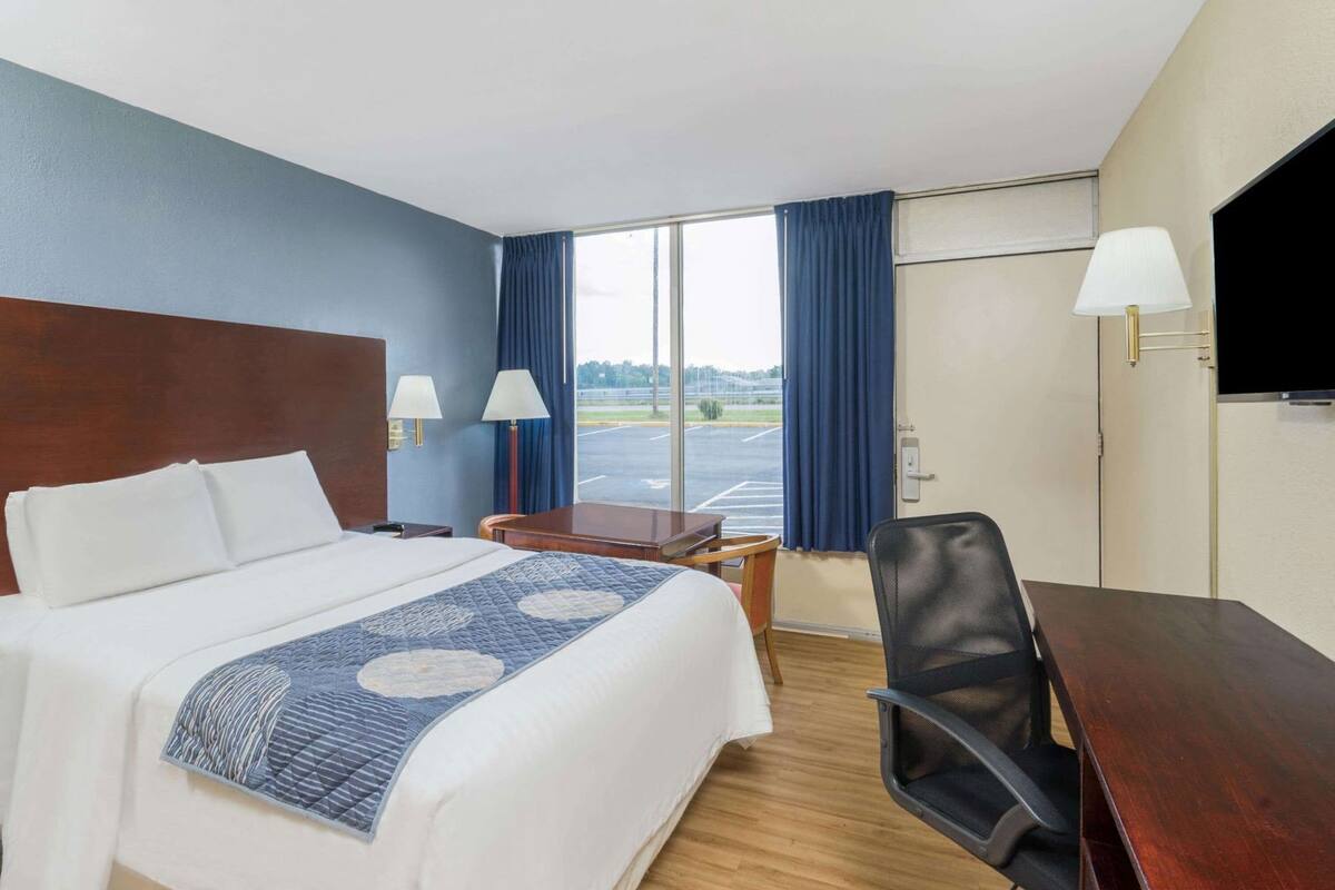 A hotel room with a bed, desk, chair, TV, and a view of a parking lot.