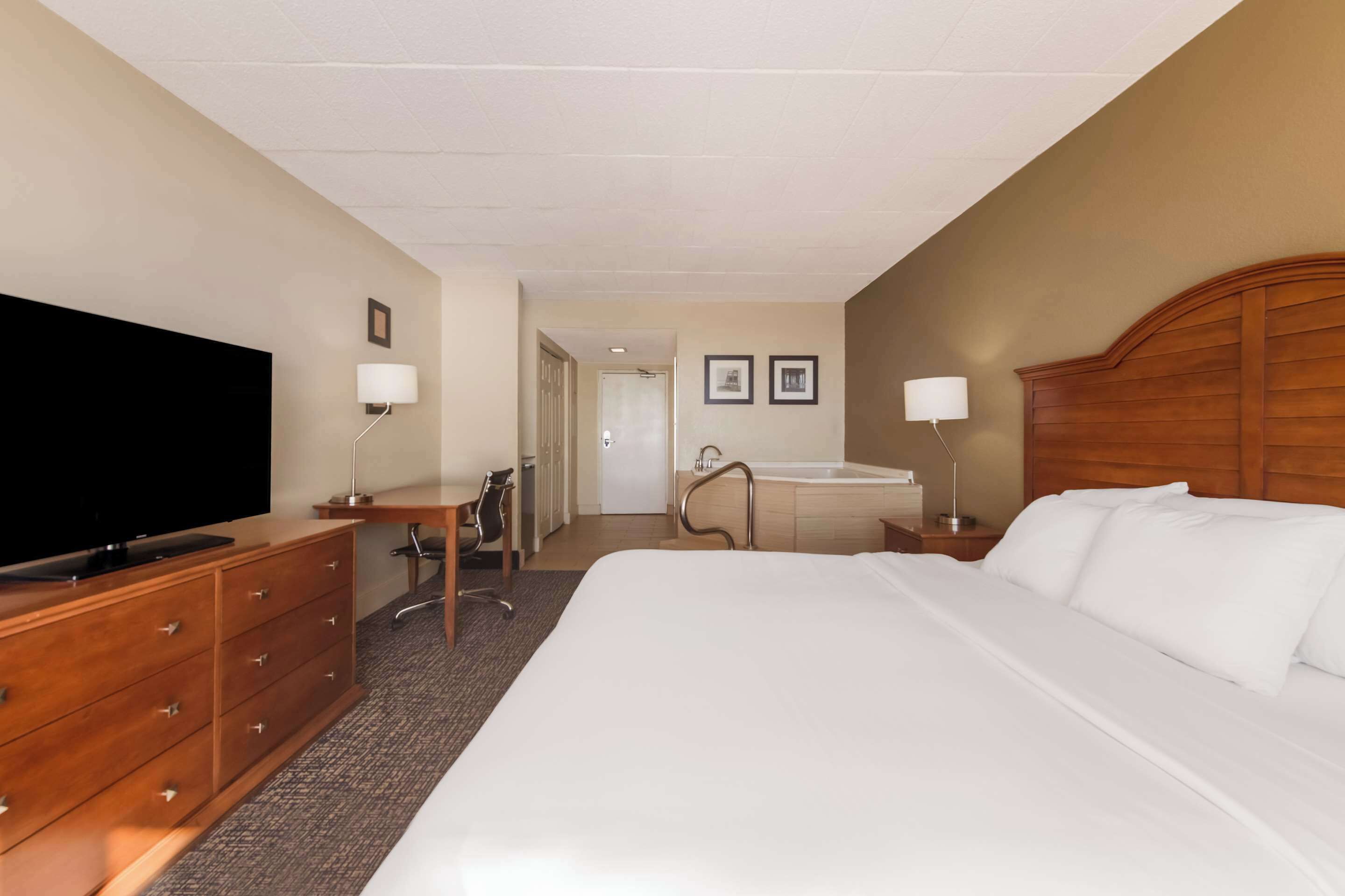 A hotel room with a large bed, a desk with a chair, a television, a bathroom with a bathtub, and a wooden headboard.