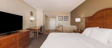 A hotel room with a large bed, a desk with a chair, a television, a bathroom with a bathtub, and a wooden headboard.