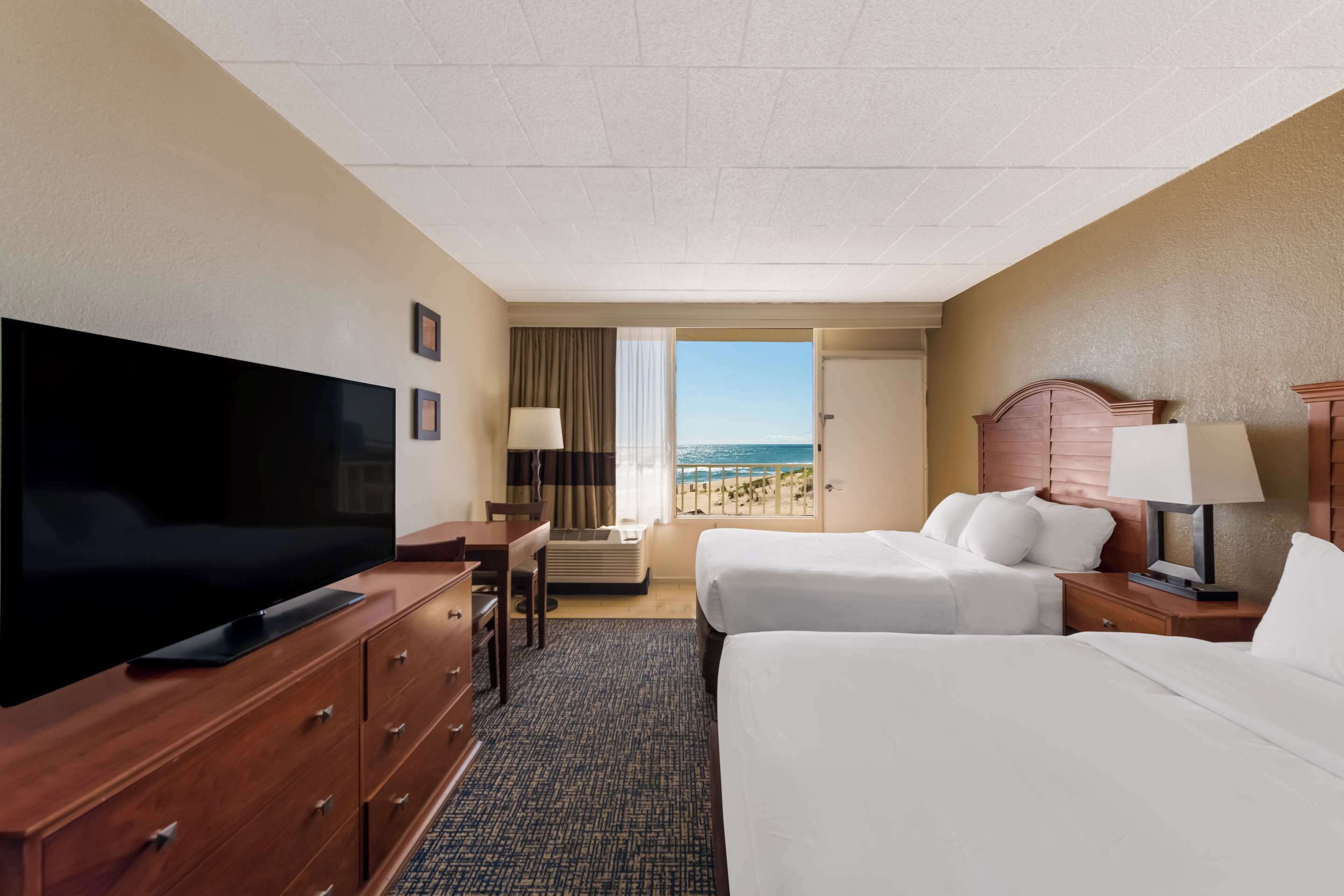 A hotel room with a TV, two beds, a desk, and a view of the beach.