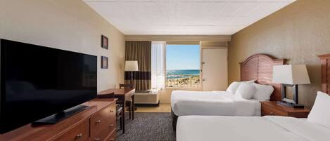 A hotel room with a TV, two beds, a desk, and a view of the beach.