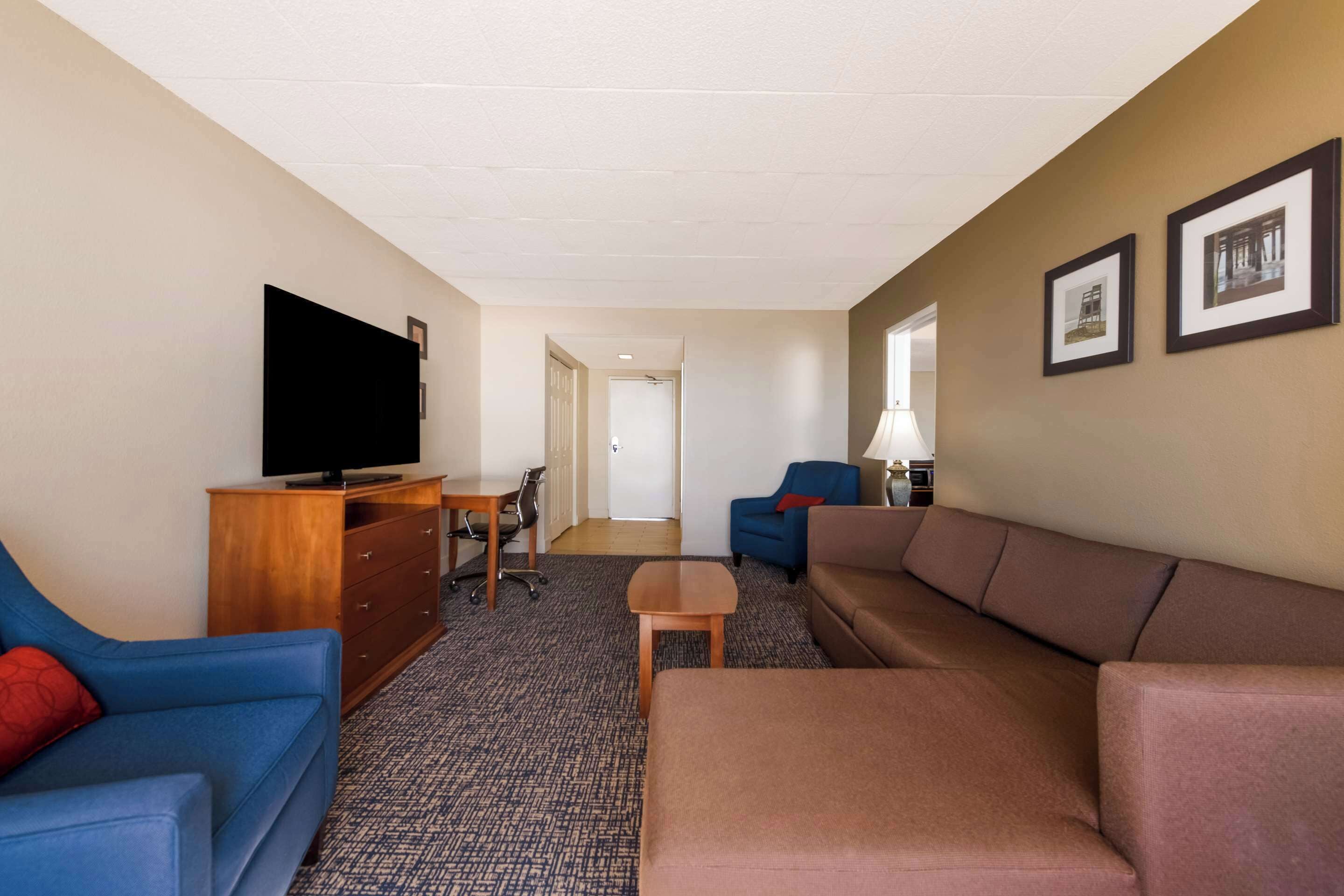 A hotel room with a large flat-screen TV, a blue sofa, a brown sofa, a wooden dresser, and a small table.