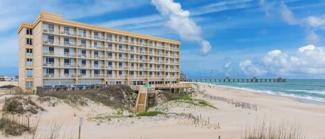 A multi-story hotel with balconies, located on a beach with a pier in the background.