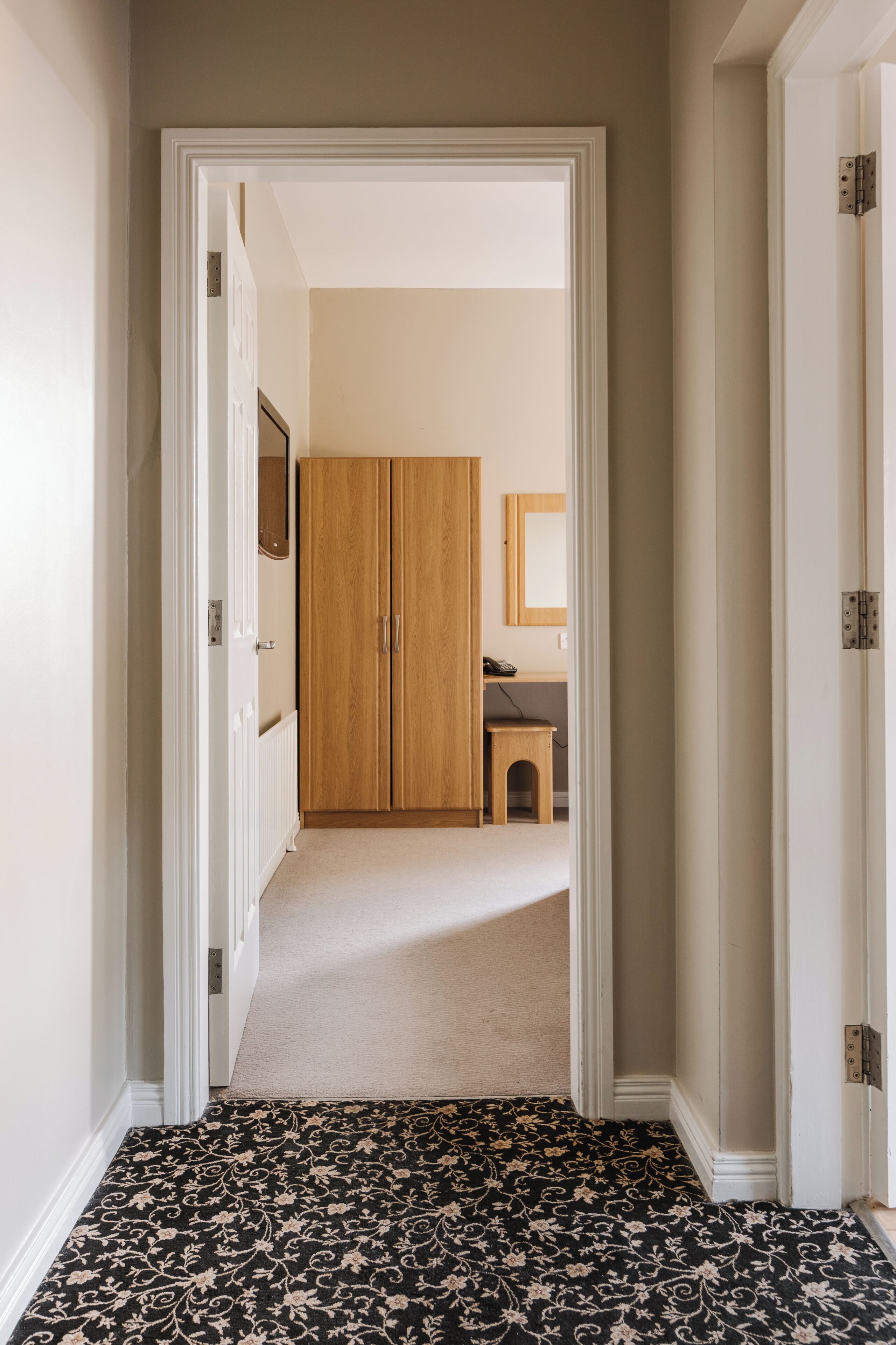 A hallway with a carpeted floor leading to a room with a wooden wardrobe and a small bench.