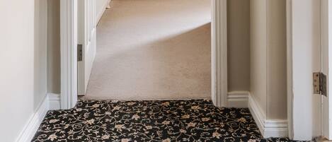 A hallway with a carpeted floor leading to a room with a wooden wardrobe and a small bench.