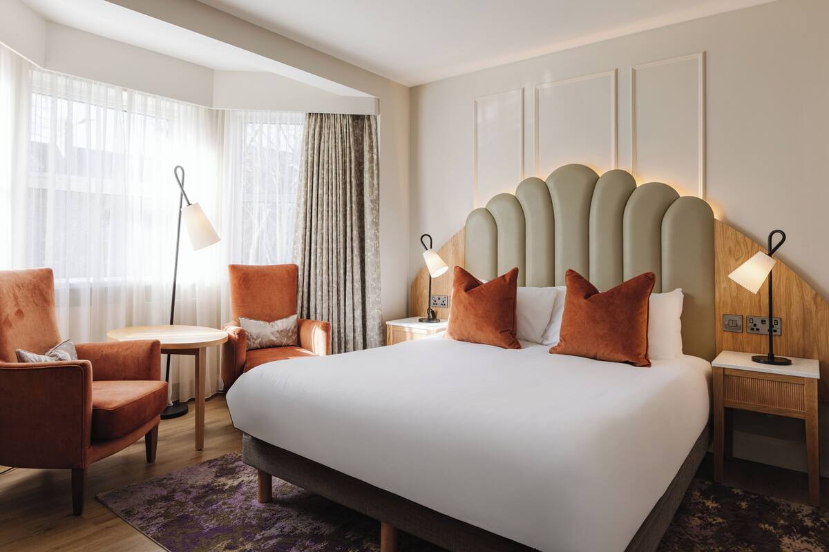 A hotel room with a large bed, two side tables, a chair, a small table, and a lamp.