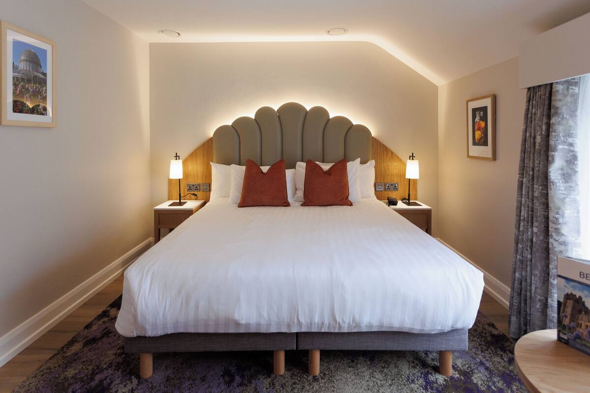 A large bed with a unique headboard, two bedside tables with lamps, a purple rug, and a framed picture on the wall.