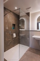 Un baño moderno con ducha envuelta en cristal, paredes de mármol, un lavabo blanco y un espejo pequeño.