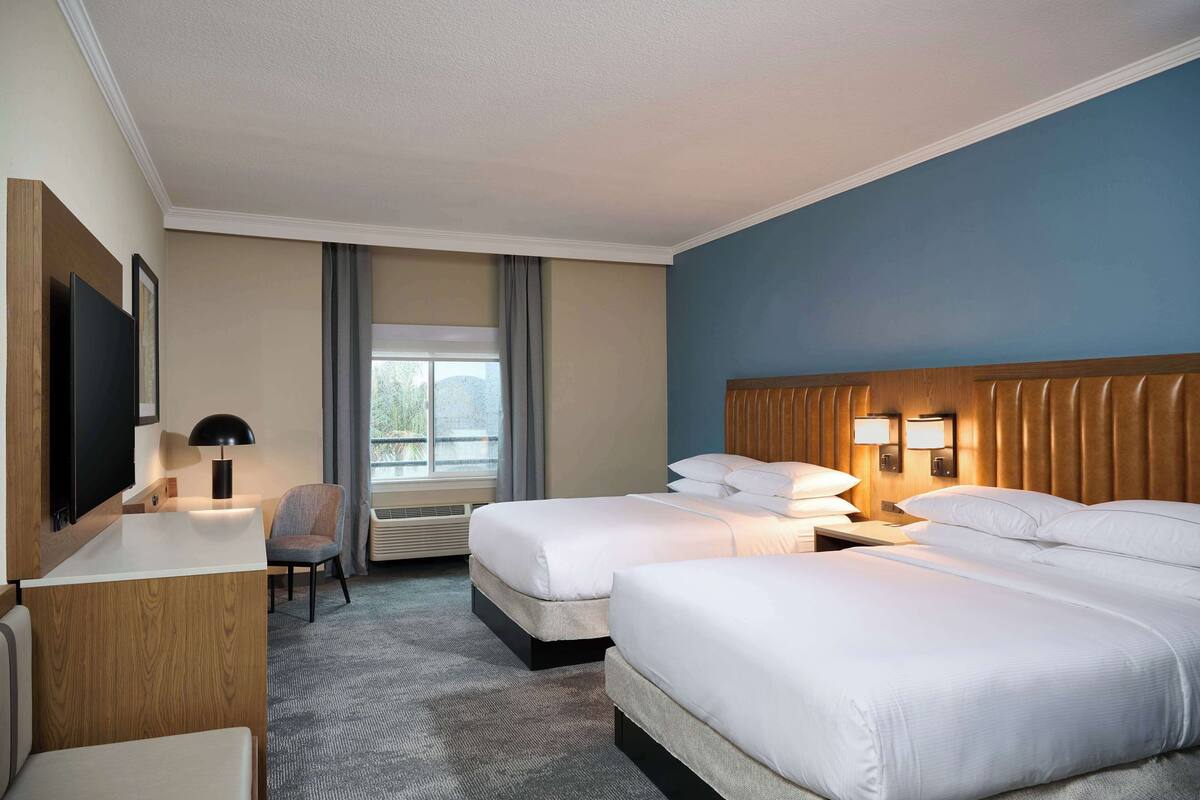 A hotel room with two beds, a desk, a chair, a TV, and a window with curtains.