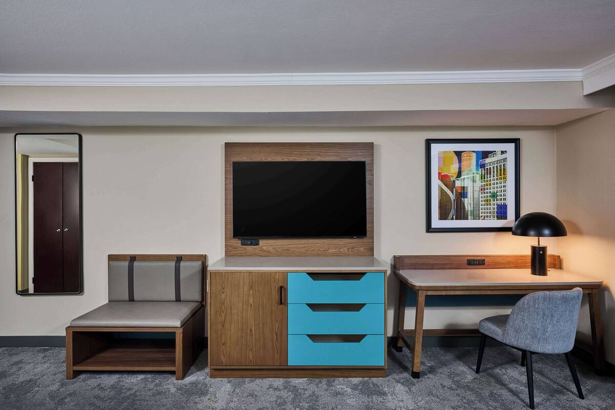 A hotel room with a wooden desk, a blue drawer unit, a grey chair, a flat-screen TV mounted on the wall, and a framed picture above the TV.