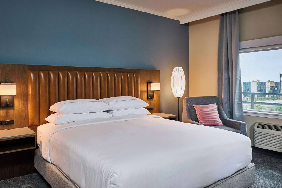 A hotel room with a large bed, two bedside tables with lamps, a chair with a pillow, and a window with curtains.