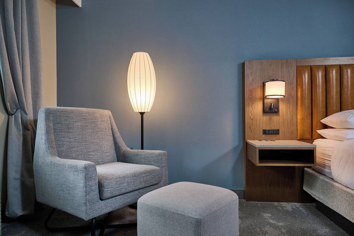 A hotel room with a grey armchair, a bedside table with a lamp, and a bed with white linens.