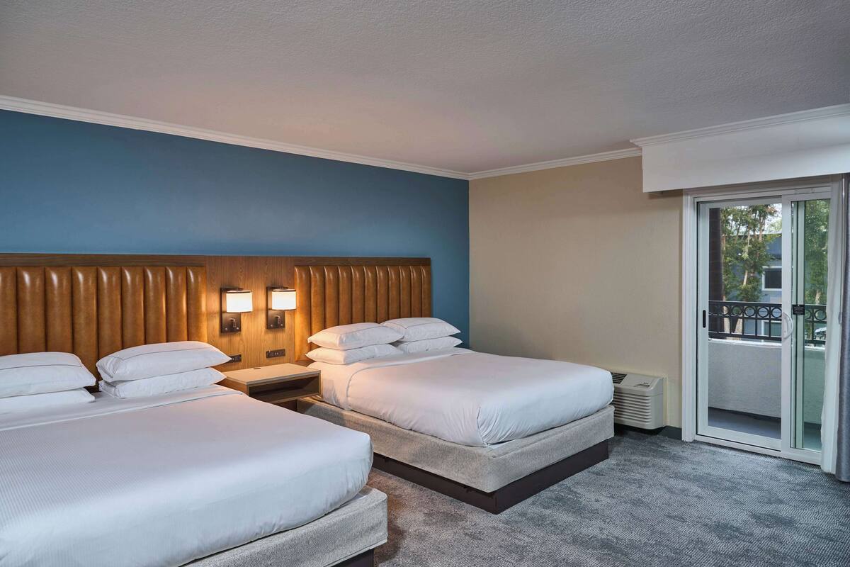 A hotel room with two beds, a wooden headboard, a nightstand with lamps, and a sliding glass door to a balcony.
